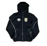 Campera deportiva negra con capucha, cierre frontal metálico y detalles de ribetes blancos. Presenta un escudo bordado en el pecho con motivos uruguayos y un logo reflectivo circular en el lado opuesto. Cuenta con bolsillos laterales con cierre.