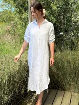 Vestido camisero largo de lino blanco, con cuello camisero, manga corta enrollada y cierre frontal de botones de nácar. Presenta aberturas laterales en el bajo.