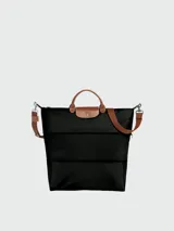 Bolso de viaje Longchamp modelo Le Pliage Original, de diseño ampliable, confeccionado en nylon negro con detalles en cuero marrón en las asas y solapa frontal con cierre de botón a presión. Incluye correa ajustable y extraíble.