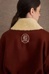 Campera tipo bomber de paño color marrón, con cierre frontal, puños y ruedo elásticos. Presenta un cuello de piel sintética desmontable en color crema y el logo de la marca bordado en el pecho.