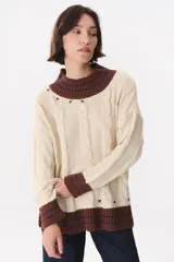 Buzo de tejido de punto con diseño de trenzas, color crema con detalles en marrón en cuello, puños y ruedo. Presenta líneas finas en contraste y pequeñas costuras decorativas en forma de cruz.