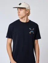 Remera blanca de algodón orgánico con cuello redondo y manga corta. Presenta un estampado en el pecho con una cruz formada por dos tablas de surf en tonos rojo, azul y negro.