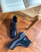 Botas cortas negras con plataforma dentada, confeccionadas en una combinación de cuero con textura de reptil y paneles de gamuza. Presentan un cierre frontal metálico y puntera cuadrada.