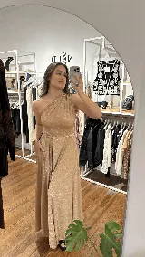 Vestido largo de fiesta color dorado con acabado brillante, diseño de un solo hombro y falda amplia con abertura lateral. Incluye un lazo ajustable en la cintura para entallar la silueta.