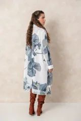 Vestido tipo túnica de corte irregular, con cierre frontal de botones y cuello camisero. Presenta un estampado botánico en tonos azules y verdes sobre fondo blanco, con detalles bordados brillantes en la parte frontal.