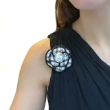 Broche en forma de flor camelia, confeccionado en tela con estampado de cuadros tipo pied-de-poule en blanco y negro, con una perla central y bordes definidos. Incluye un gancho versátil para sujetar en prendas o en el cabello.