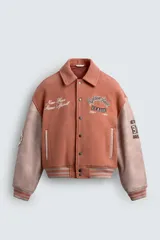 Cazadora estilo varsity con cuerpo de paño color salmón y mangas de cuero sintético en tono rosado. Presenta cuello solapa, cierre frontal con botones a presión, bolsillos laterales de vivo y puños y ruedo acanalados. Incluye parches y bordados decorativos en el frente y las mangas.
