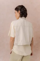 Chaleco de jean color crudo, con calce relajado y largo a la cintura. Cuenta con cuello camisero, cierre frontal con botones metálicos y bolsillos delanteros con tapa.