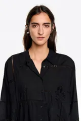 Camisa de corte recto en color negro, con cuello camisero, cierre frontal de botones y manga larga con puños abotonados. Presenta un bolsillo de plastrón en el pecho y detalles de bordados a tono con aplicaciones de encaje tipo entredós distribuidos por toda la prenda.