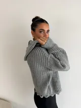 Buzo de punto grueso color gris, con cuello polo, cierre frontal parcial y capucha. Presenta un diseño de tejido acanalado con terminaciones desflecadas en el ruedo.