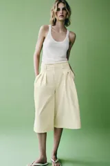 Bermudas de tiro medio color amarillo claro, confeccionadas en mezcla de algodón. Presentan cintura con trabillas, bolsillos laterales ocultos en costura, pliegues delanteros y cierre frontal con cremallera, botón y ganchos metálicos.