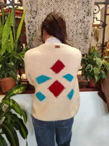 Cardigan de lana merino tejido a mano con técnica de dos agujas, de color marfil con parches geométricos de tela en tonos celestes y lila aplicados con costuras visibles. Presenta cierre frontal con botones de madera y detalles de ribetes multicolores en los bordes.