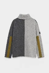 Polera de tejido lambswool con diseño bicolor en tonos neutros. Presenta cuello alto tejido, mangas largas con franjas laterales en color marrón y detalle de bordado artesanal con tres pequeñas líneas horizontales en colores vibrantes.