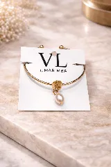 Pulsera rígida abierta de tono dorado con un dije central con forma de caracola marina y una perla colgante de forma ovalada.