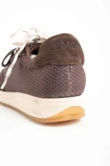 Zapatilla deportiva de perfil bajo, color marrón chocolate, con paneles de gamuza y malla texturizada. Presenta una entresuela de color crema y suela de goma color caramelo. Se ata con dos pares de cordones: uno blanco trenzado y otro verde oscuro.