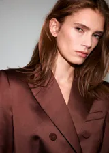 Blazer de corte recto y diseño estructurado, confeccionado en tejido satinado color marrón. Presenta cuello con solapas de muesca, cierre frontal de doble botonadura, bolsillos de solapa en el frente y bolsillo de parche en el pecho. Incluye forro interior y mangas largas con puños abotonados.