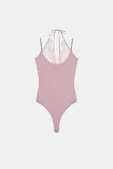 Body de tejido elástico color rosado, con escote redondo, tirantes finos y detalle de encaje en el frente con lazada ajustable al cuello. Cuenta con cierre inferior mediante botones a presión.