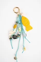 Charm para bolso con argolla dorada, cordones de hilo celeste y colgantes variados que incluyen cuentas de cerámica, conchas marinas, abalorios de cristal y un dije grande en forma de pez de color amarillo.