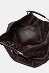 Bolso tipo saca de piel color marrón oscuro, con cuerpo fruncido y doble asa de mano rígida metálica. Incluye asa extraíble para llevar al hombro o cruzado y bolsillo interior extraíble.