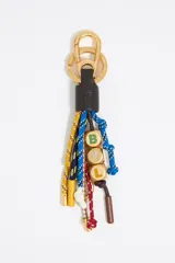 Llavero de la marca Bimba y Lola con herrajes metálicos dorados y múltiples cabos trenzados de colores. Presenta una pieza central de cuero negro con el logo grabado y cuentas cúbicas doradas con las iniciales 'B', 'Y' y 'L' en relieve. Los cordones incluyen diversos nudos y terminaciones metálicas decorativas.