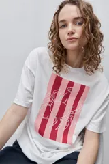 Remera blanca de calce relajado y silueta amplia, con cuello redondo, mangas cortas y estampa gráfica frontal de un cangrejo sobre franjas en tonos rosado y rojo.