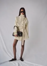 Chaqueta de algodón elastizado color beige, con cuello alto, cierre frontal de botonadura simple, mangas largas y cinturón ajustable del mismo tejido.