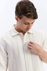 Camisa de manga corta de corte oversize en tejido rústico con diseño de rayas verticales celestes sobre fondo beige, cuello clásico y cierre frontal de botones.