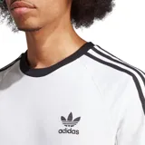 Remera blanca de algodón con cuello redondo y mangas cortas con ribete azul marino. Presenta las tres rayas características de Adidas en azul marino en los hombros y el logo de Adidas bordado en el pecho.