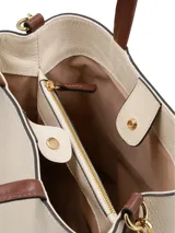 Cartera tipo tote de cuero color crudo con detalles en marrón. Tiene doble asa corta, correa larga extraíble, cierre con broche metálico, logo metálico frontal y bolsillo interno con cierre. Diseñada en Uruguay y fabricada en Brasil.