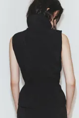 Chaleco negro de diseño fluido con cuello subido y cierre frontal de cremallera metálica. Presenta cordones ajustables con lazadas en el cuello y en la cintura.