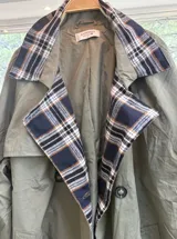 Gabardina de corte clásico en color verde militar, con solapas amplias y puños con detalles en estampado de cuadros escoceses en tonos azul, blanco y negro. Presenta cierre frontal cruzado con botones y bolsillos laterales.