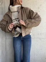 Campera bomber de gamuza color beige, con cierre frontal y detalle de ojales metálicos decorativos en la solapa. Presenta mangas largas con puños elásticos y un corte holgado tipo oversize.