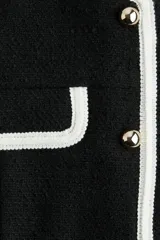 Chaleco corto negro en tejido bouclé, con cuello redondo, cierre frontal de botones dorados y bolsillos frontales decorativos ribeteados en blanco.