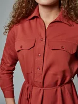 Vestido camisero color verde óxido, con cuello camisero, mangas largas con puños abotonados, dos bolsillos con solapa en el pecho, cinturón de lazo y cierre frontal con botones.