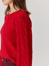 Buzo de lana color beige con mangas abullonadas de tejido calado con diseño de rombos. Presenta un cuello redondo y un detalle de flor negra de tela con cintas colgantes en el escote.