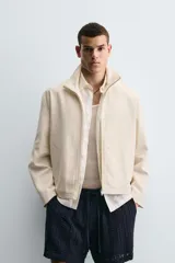 Cazadora de tejido color crema, con cuello alto, cierre frontal de cremallera y bolsillos de vivo en la cadera. Presenta un calce relajado (relaxed fit).