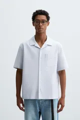 Camisa de algodón con rayas verticales finas azules y blancas, corte relajado (relaxed fit), cuello bowling y manga corta. Posee un bolsillo plastrón en el pecho y aberturas laterales en el bajo.
