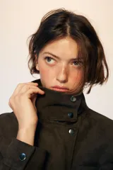 Cazadora tipo bomber de tejido efecto ante color marrón oscuro, con cuello subido, manga larga y cierre frontal con cremallera y botones ocultos. Presenta bolsillos frontales con solapa y botones metálicos.