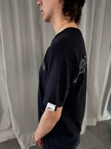 Remera de algodón color negro con estampado serigráfico en tono gris oscuro en la parte posterior. Presenta cuello reforzado, costuras reforzadas y una pequeña etiqueta decorativa en la manga.
