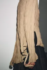 Top de manga larga color beige, con textura fruncida en todo el cuerpo y mangas. Presenta cuello redondo y bajo asimétrico con aberturas laterales ajustables con cordones.