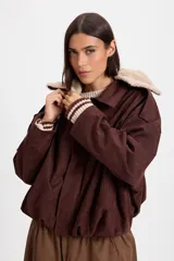 Campera bomber de gamuza color chocolate con cuello de corderito sintético desmontable en color crema. Presenta un diseño holgado con cierre frontal, hombreras decorativas y bordado sutil en el pecho.