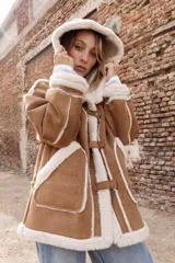 Campera estilo gamulán color tostado con interior y bordes de corderito sintético color crema. Presenta capucha amplia, cierre frontal con trabas y broches metálicos, y bolsillos delanteros grandes. Posee un calce relajado y diseño de textura suave.