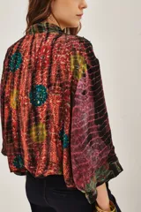Kimono de estilo abierto con mangas anchas, confeccionado en viscosa con un diseño estampado que combina patrones tipo tie-dye y patchwork en tonos vibrantes. Presenta detalles de aplicaciones decorativas y forro interior.