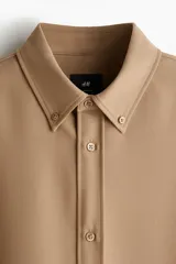 Camisa de manga larga color camel, confeccionada en sarga suave, con cuello americano y tapeta clásica con botones. Presenta canesú en la espalda y bajo redondeado.