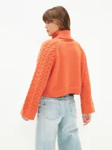 Polerón de tejido de punto color gris claro, con cuello alto y diseño de trenzas en el cuerpo y mangas. Corte holgado y puños anchos.