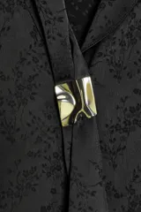 Camisa de satén negro con diseño jacquard de flores tono sobre tono. Presenta escote en pico con solapas, cierre frontal con botones y un broche metálico decorativo en el centro. Tiene mangas largas con puños abotonados.
