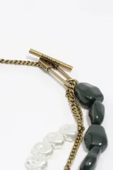 Collar de diseño asimétrico compuesto por una combinación de cadenas de eslabones plateados, una cadena de esferas metálicas doradas y una sección con cuentas de resina en tonos verde oscuro y blanco traslúcido. Presenta un cierre tipo T en la parte superior.