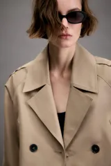 Trench corto de color beige con cuello de solapa, cierre frontal cruzado con botones y mangas largas con trabillas ajustables en los puños. Presenta un diseño estructurado con hombreras y bolsillos delanteros de vivo.
