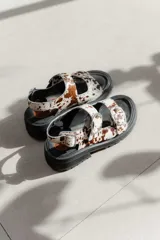 Sandalias de plataforma con diseño de dos tiras ajustables con hebillas metálicas y tira trasera con hebilla. Confeccionadas en cuero con estampado animal print tipo vaca, sobre una base de goma negra.