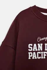 Sudadera de cuello redondo y manga larga, color bordó, con estampado frontal en blanco que dice "Chasing The Sunset's Glow SAN DIEGO 1980 PACIFIC HARBOR".